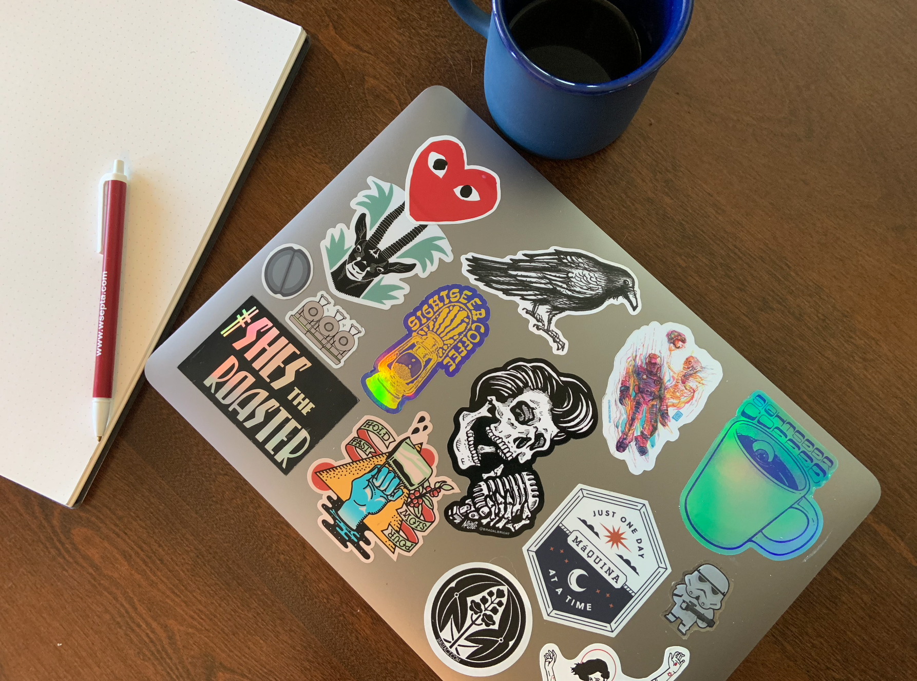 This image contains a laptop with many stickers on it, resting on a desk. The stickers on the laptop include the blue holographic Sightseer mug and purple holographic Sightseer lantern stickers.